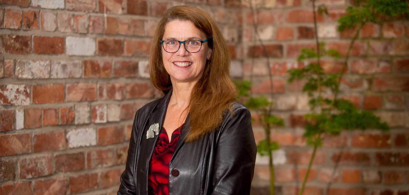 smiling woman in black top and black jacket, against a red brick wall